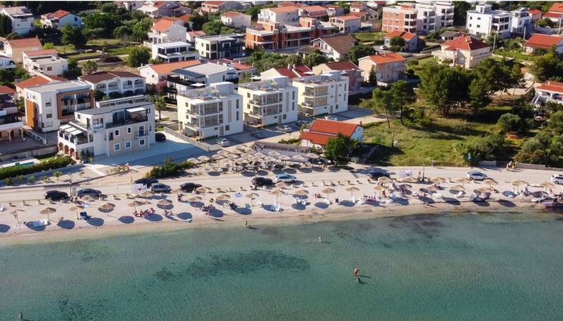 Ein Strand mit Liegestühlen und Sonnenschirmen, dahinter Häuser und Gebäude im Ferienort.