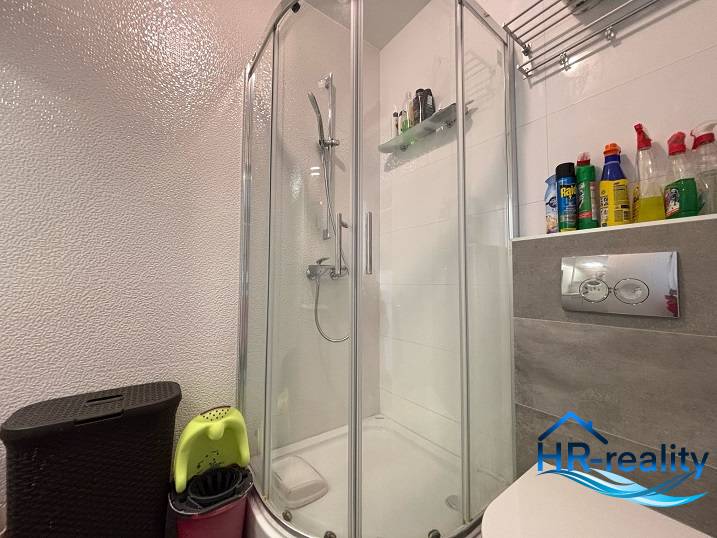 Badezimmer in einer 2-Zimmer-Wohnung mit einer Eckdusche und einem Regal für Reinigungsmittel.