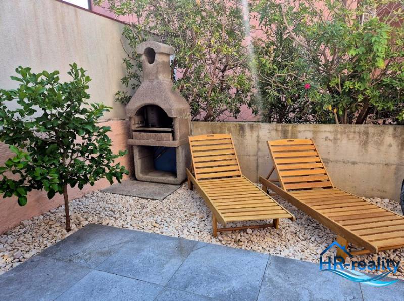 Gartenbereich einer 2-Zimmer-Wohnung in Novalja mit Liegestühlen und Grill, umgeben von Grün.