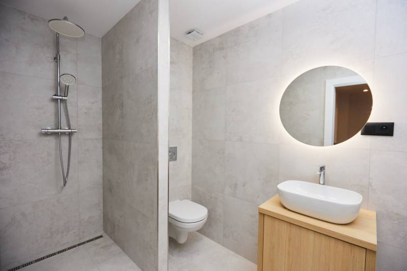 Badezimmer in einer 2-Zimmer-Wohnung mit offener Dusche und beleuchtetem Spiegel.