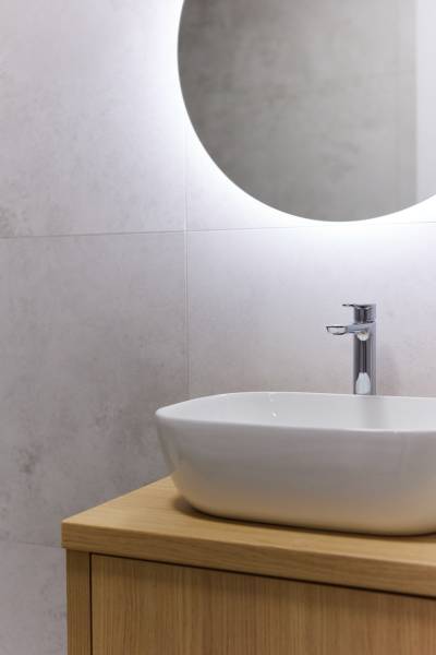 Badezimmer mit Waschbecken auf einem Holzschrank und beleuchtetem rundem Spiegel in einer Zweizimmerwohnung.