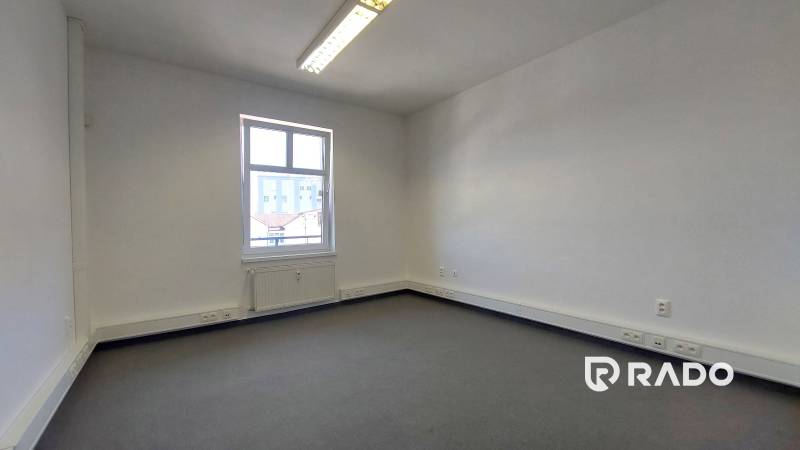 Ein leeres Büro mit einem Fenster, weißen Wänden und grauem Boden, geeignet zur Vermietung.