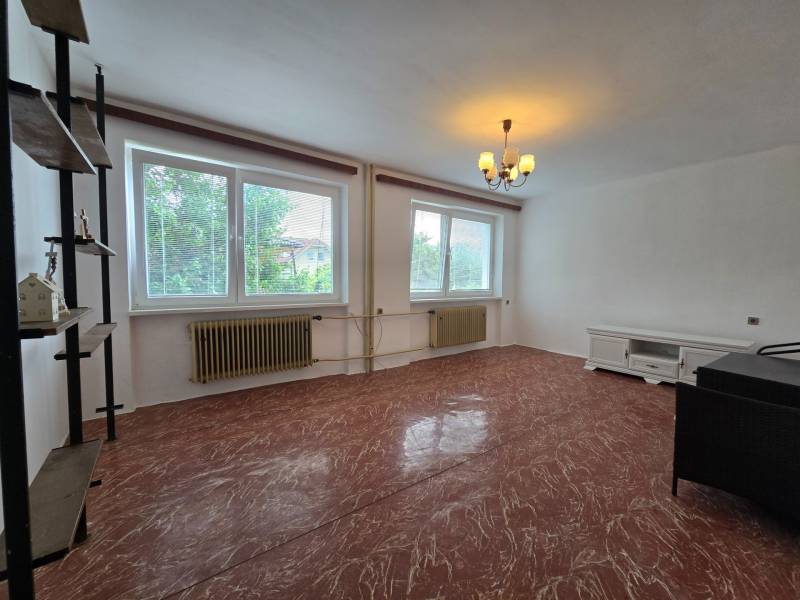 Wohnzimmer in einem Einfamilienhaus, mit weißen Wänden und rotem Boden, zwei Fenstern und Möbeln.