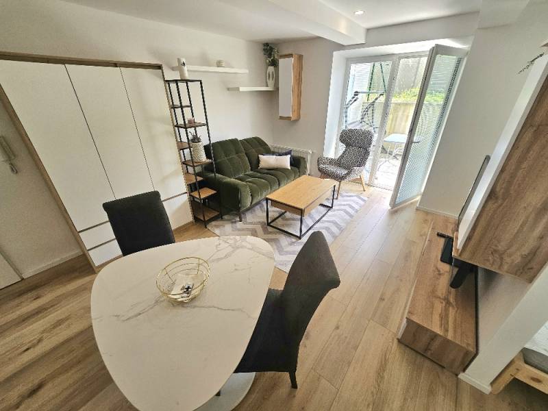 Wohnzimmer in einer Einzimmerwohnung mit grünem Sofa, Esstisch und Sessel beim Balkon.