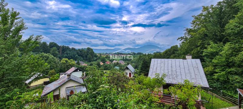Blick auf die Hütten in Mojš, umgeben von üppigem Grün und Hügeln unter dramatischem Himmel.