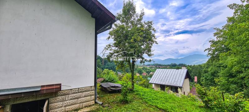 Der Blick von der Hütte in Mojš zeigt Dächer, Grünflächen und entfernte Hügel unter einem bewölkten Himmel.