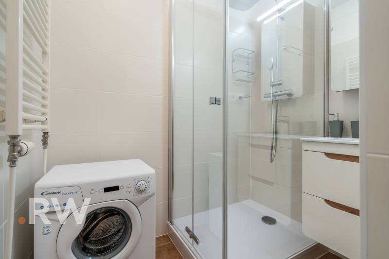Badezimmer in einer 2-Zimmer-Wohnung mit Waschmaschine, Duschkabine und weißen Fliesen.