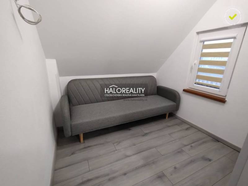 Ein Studio-Apartment mit einem grauen Sofa, weißen Wänden und einem Fenster mit Jalousien.