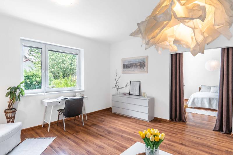Helles Wohnzimmer mit Holzboden, Schreibtisch am Fenster und Bett hinter einem Vorhang.