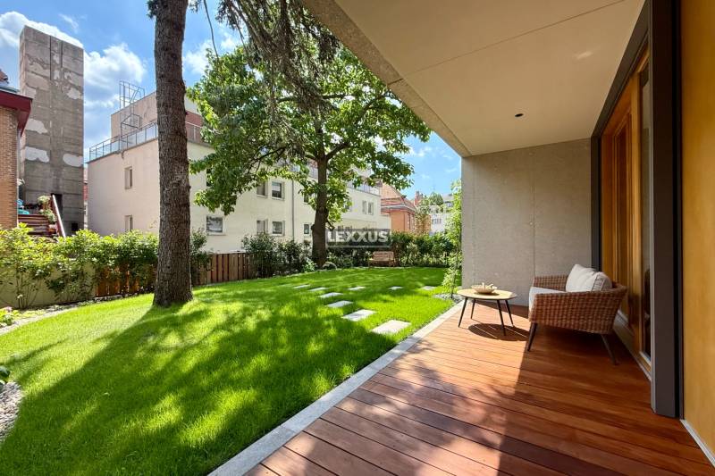 Terrasse mit Garten bei einer 4-Zimmer-Wohnung, Bäume und Gebäude im Hintergrund.