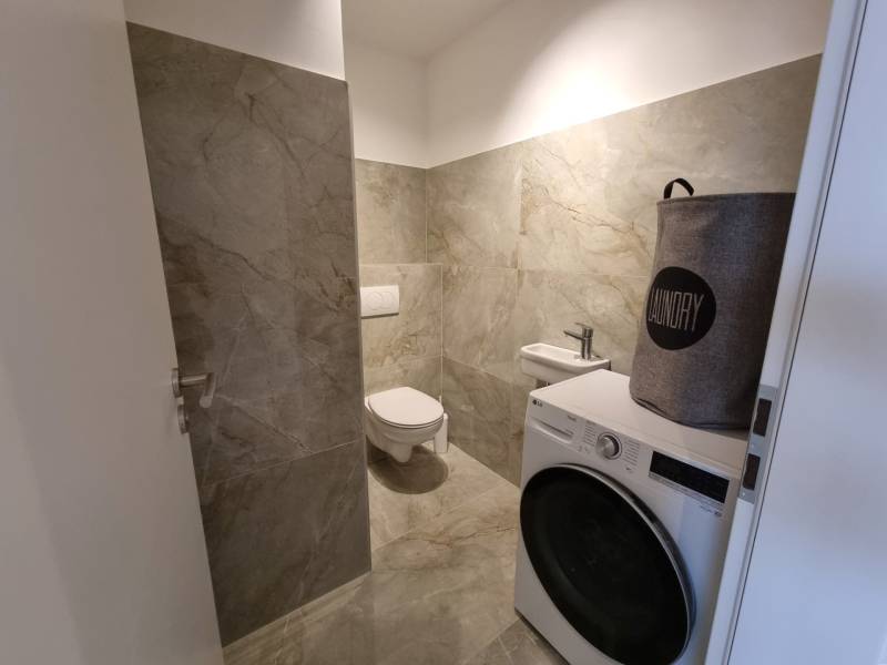 Badezimmer mit grauen Fliesen, Waschmaschine und Wäschekorb in einer 3-Zimmer-Wohnung.