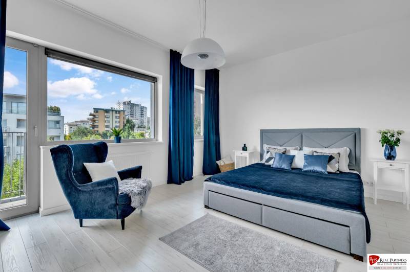 Helles Schlafzimmer in einer 4-Zimmer-Wohnung mit großem Fenster, blauen Vorhängen und einem bequemen Sessel.