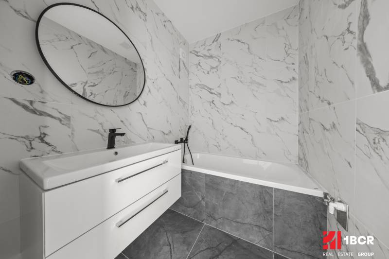 Badezimmer in einer 2-Zimmer-Wohnung mit Badewanne, weißem Waschbecken und rundem Spiegel.