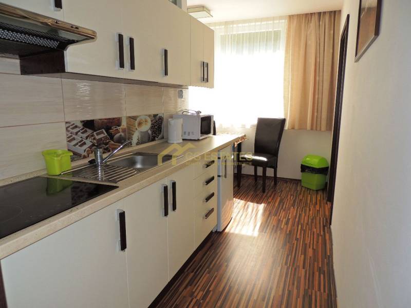 Küche in einer 2-Zimmer-Wohnung mit weißen Schränken, Holzboden und einem Fenster mit Rollläden.
