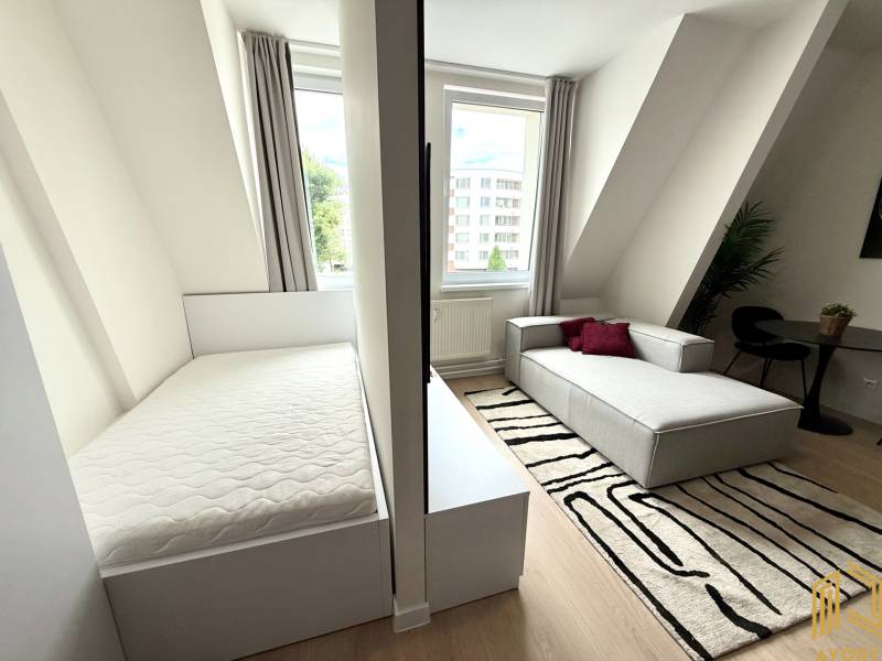 Garsónka mit einem hellen Bett, einem grauen Sofa und einem schwarz-weißen Teppich.