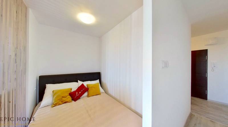 Schlafzimmer mit Doppelbett, weißen Wänden und bunten Kissen in einer Einzimmerwohnung.