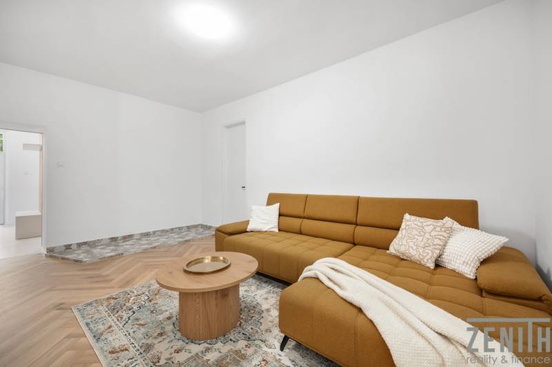 Ein Zimmer in einer 3-Zimmer-Wohnung mit einem braunen Sofa, einem Tisch und einem gemusterten Teppich.