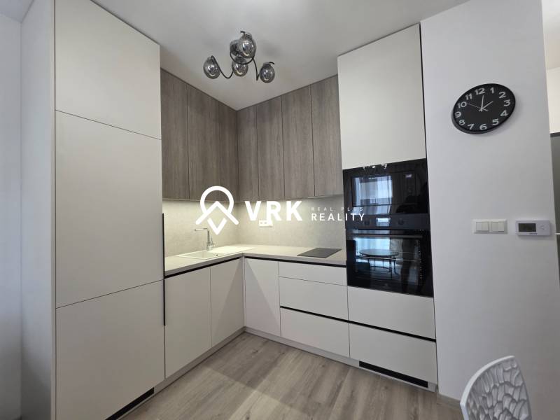 Küche in einer 2-Zimmer-Wohnung mit weißen und hölzernen Schränken, eingebauten Geräten, Uhr.