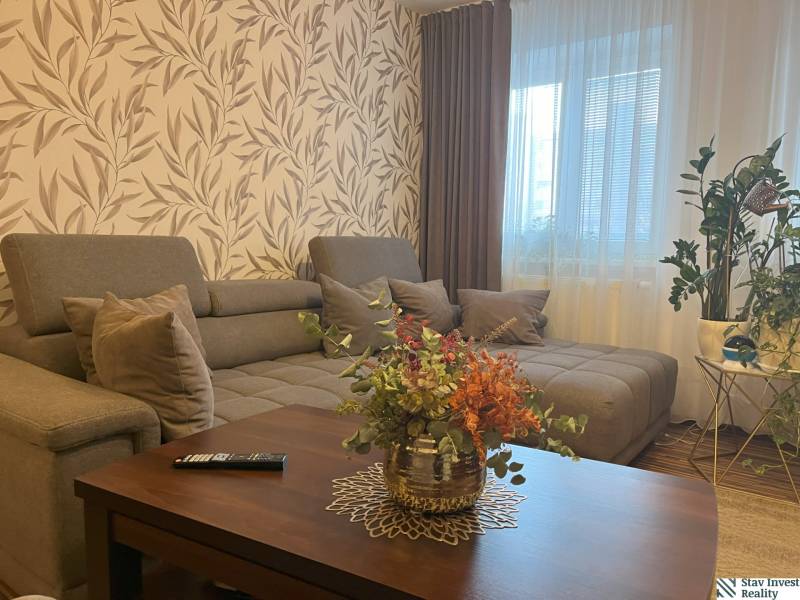 Wohnzimmer einer Zweizimmerwohnung mit großem Sofa, Blumen und gemusterten Tapeten.