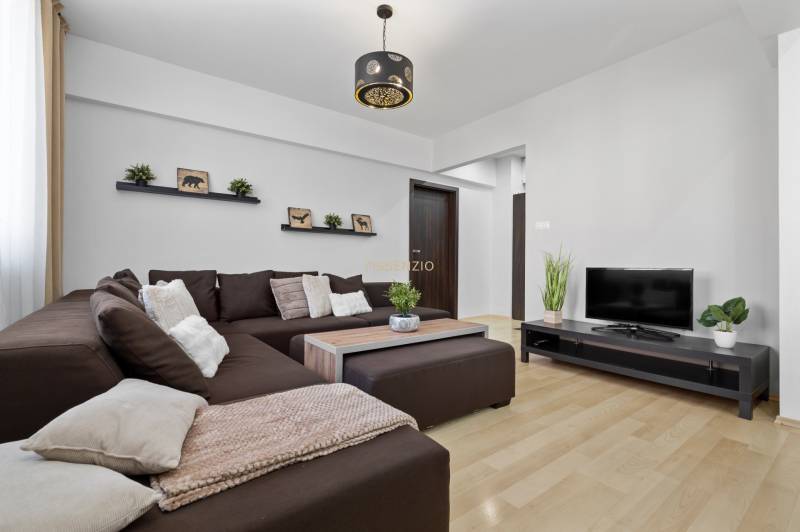 Wohnzimmer in einer Zweizimmerwohnung mit braunem Sofa, Fernseher und Boden in Holzoptik.