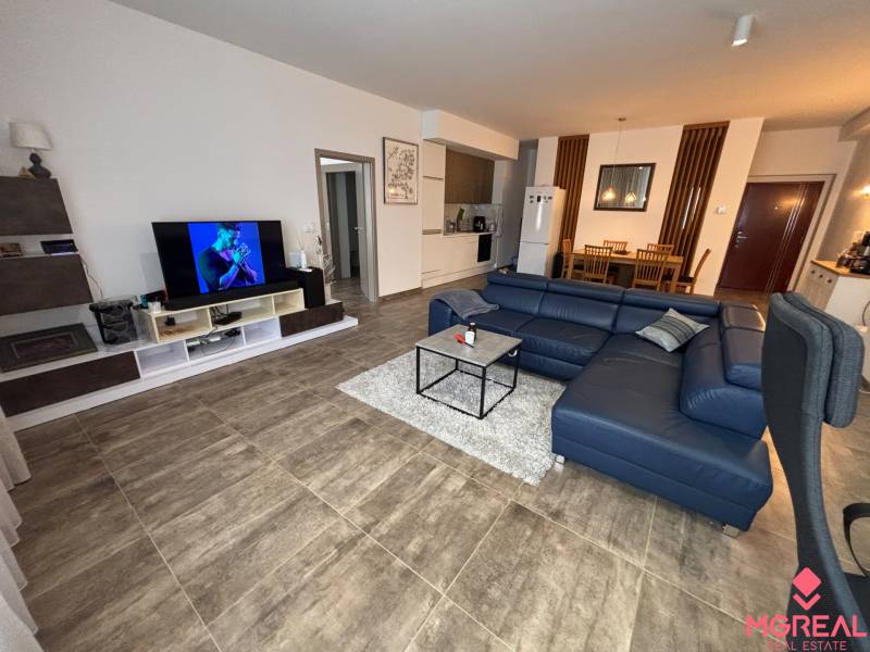 Wohnzimmer in einer Zweizimmerwohnung mit dunklem Sofa und Fernseher, Boden mit Holzdekor.