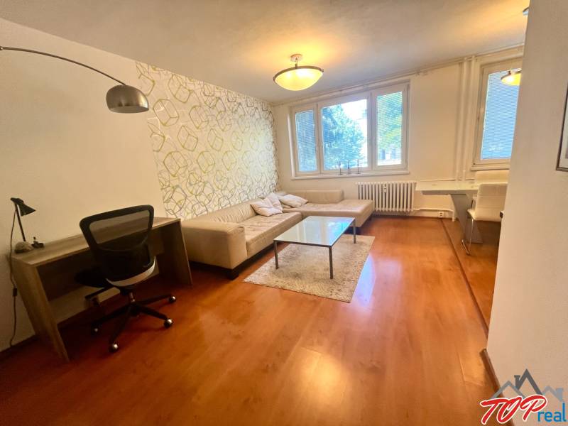 Obývací pokoj v třípokojovém bytě s pohovkou, stolkem a pracovním stolem.