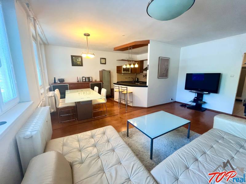 Wohnzimmer mit Küche in einer 3-Zimmer-Wohnung, Ausstattung umfasst ein Sofa, einen Tisch und einen Fernseher.