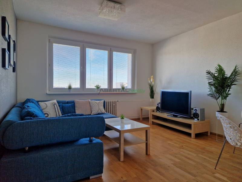 Wohnzimmer in einer 3-Zimmer-Wohnung mit einem blauen Sofa, einem Fernseher und einem Boden in Holzoptik.