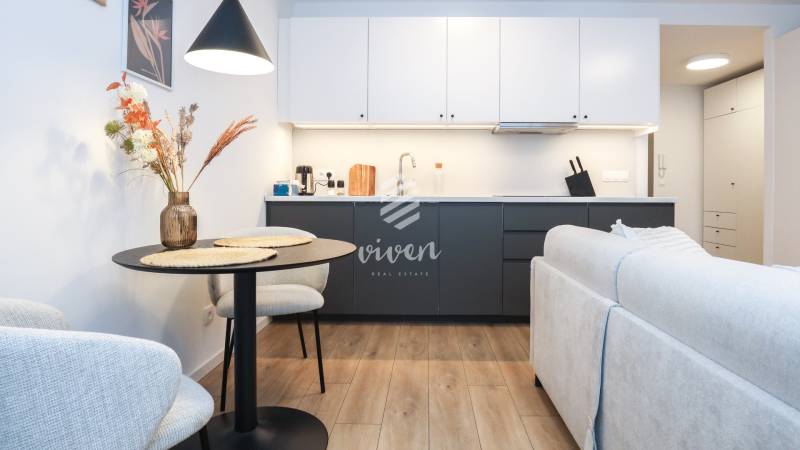 Küche in einer Einzimmerwohnung mit weißen Schränken, dunkler Arbeitsplatte und einem Boden in Holzoptik.