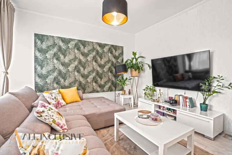 Helles Wohnzimmer mit Sofa und Blumenkissen, grüne Tapete, Fernseher an der Wand.