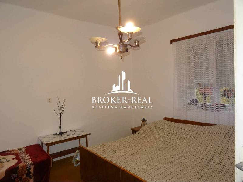 Schlafzimmer im Einfamilienhaus mit Bett, Tisch und Kronleuchter, an der Wand ist ein Fenster mit Gardine.