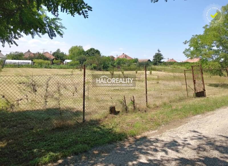 Ein ebenes Grundstück, das von einem Zaun in den Gärten von Marcelová umgeben ist, im Hintergrund sind Häuser zu sehen.