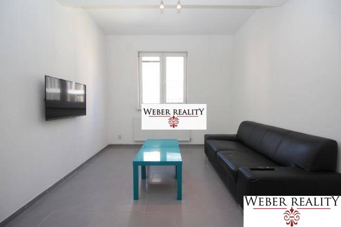 Wohnzimmer einer Zweizimmerwohnung mit schwarzem Sofa, Fernseher an der Wand und niedrigem Tisch.