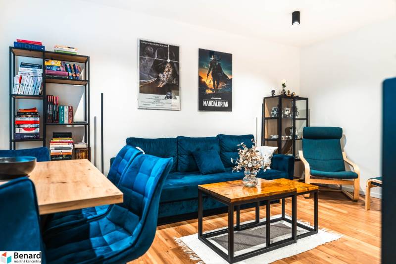 Wohnzimmer einer Zweizimmerwohnung mit einem blauen Sofa, einem Bücherregal und einem Boden in Holzoptik.