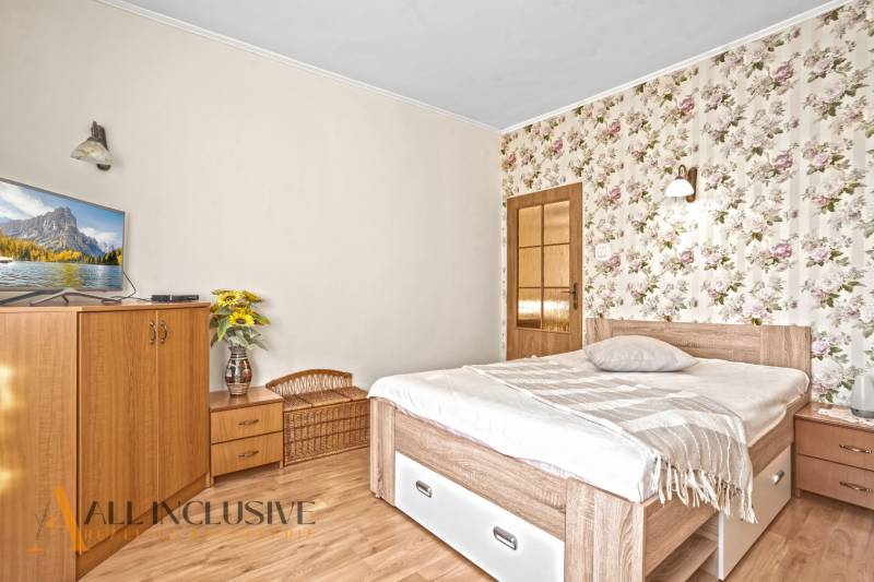 Ein Schlafzimmer in einem Einfamilienhaus mit einer Blumentapete, einem Fernseher und einem Boden in Holzoptik.