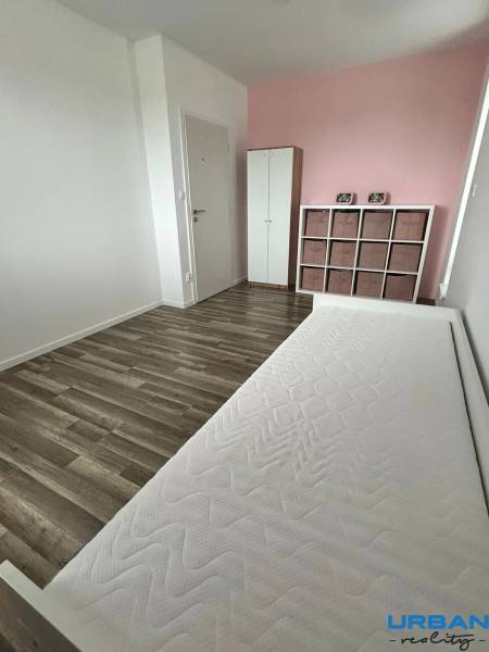 Ein helles Zimmer mit einer rosa Wand, einem Bett und einer Kommode, Boden mit Holzdekor.