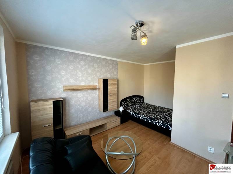 Ein Zimmer in einer Einzimmerwohnung mit einem Bett, einem schwarzen Sessel und einem Boden in Holzoptik.