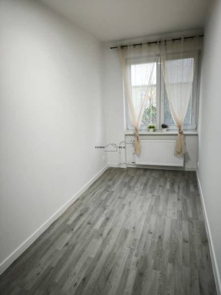 Ein kleiner Raum mit einem Holzboden, weißen Wänden und einem Fenster mit Vorhängen.