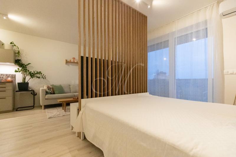 Küche mit hellen Möbeln, Sofa und Bett in einer Einzimmerwohnung, Boden mit Holzdekor.