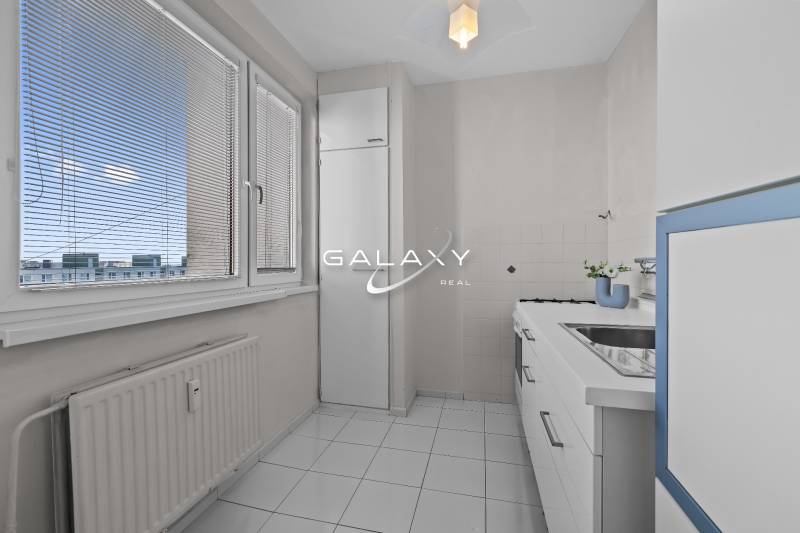 Küche in einer Einzimmerwohnung mit weißen Möbeln und Fliesen, mit Blick durch Jalousien.