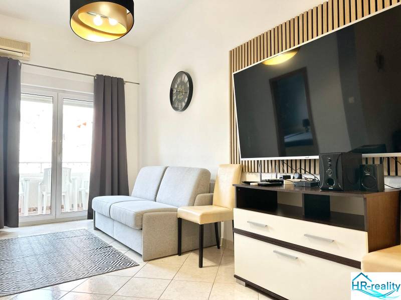 Wohnzimmer mit Sofa und Fernseher in einer Ferienwohnung, heller Fliesenboden, Balkontüren.