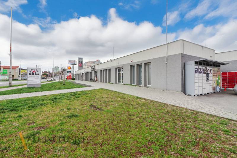 An der Vajanského-Straße befinden sich Geschäftsräume, neben dem Gehweg ist eine grüne Rasenfläche.