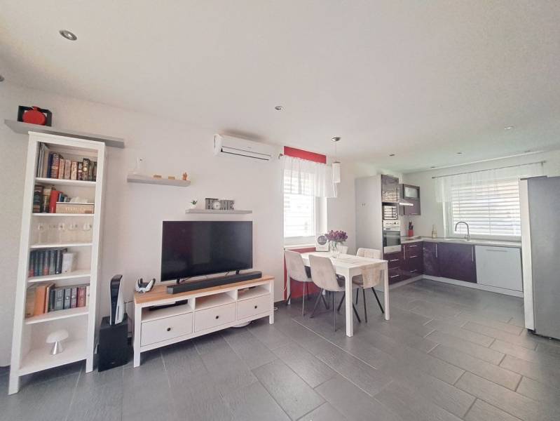 Wohnzimmer in einem Einfamilienhaus mit Fernseher, Kochnische und einem Tisch mit drei Stühlen.