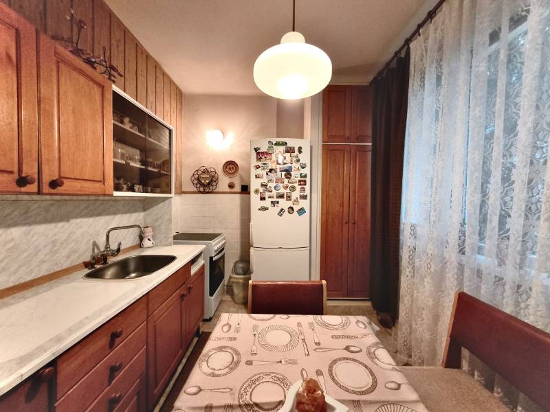 Küche in einer 3-Zimmer-Wohnung mit Holzschränken und Kühlschrank, dekoriert mit Vorhängen.