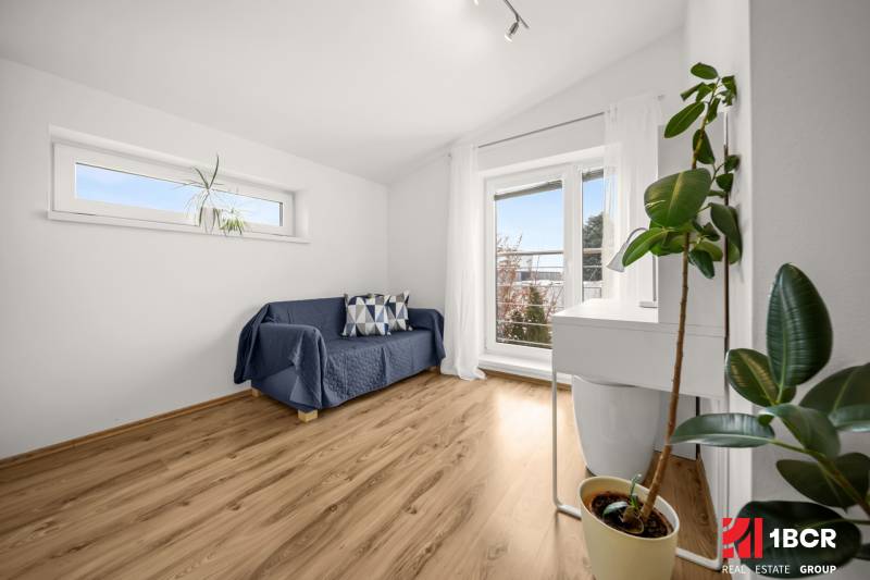 Ein Zimmer in einem Einfamilienhaus mit einem Sofa, Fenstern und einem Boden in Holzoptik.