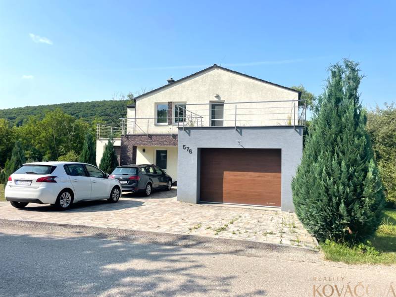 Ein Einfamilienhaus in Melčice-Lieskové, vor dem zwei Autos parken, umgeben von Grün.