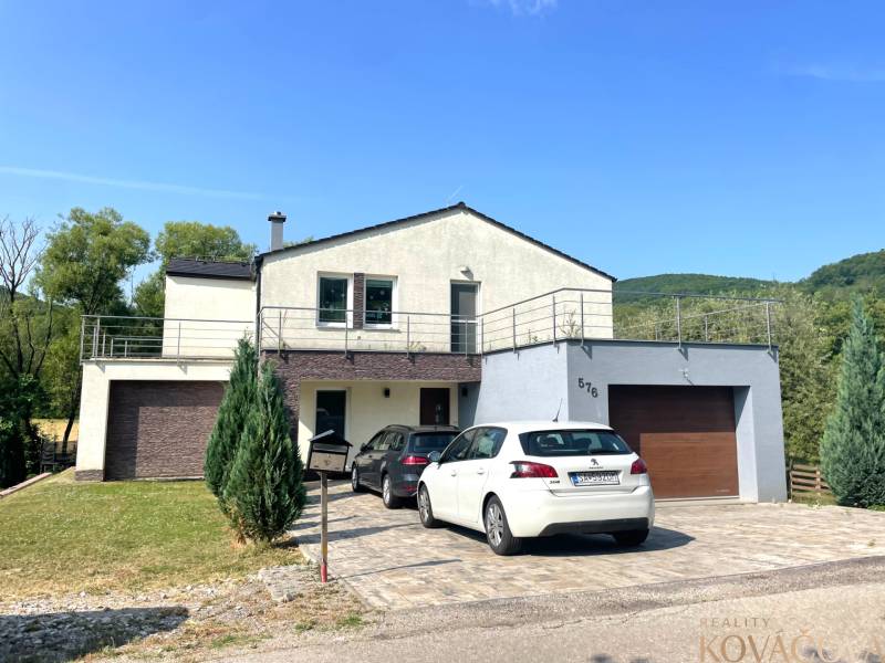 Ein Einfamilienhaus in Melčice-Lieskové mit einer Garage, zwei Autos und einem Garten auf einem Hanggrundstück.