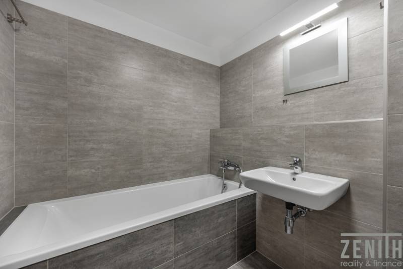 Badezimmer in einer 2-Zimmer-Wohnung mit Badewanne, Waschbecken und grauen Fliesen.