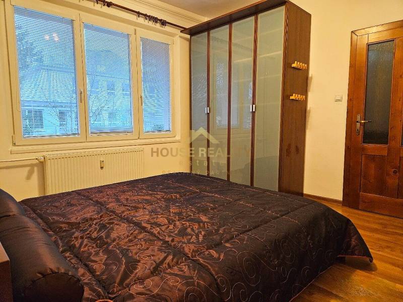 Schlafzimmer mit großem Kleiderschrank und Bett mit dunklem gestepptem Überwurf, Boden mit Holzdekor.