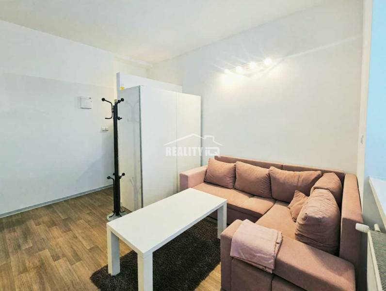 Einzimmerwohnung mit Ecksofa, weißem Tisch und Boden in Holzoptik.